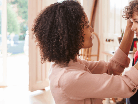 Une mère ajuste délicatement le sac d’école de son jeune fils dans l’entrée ensoleillée de la maison avant le retour en classe.