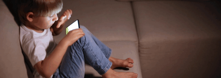 Jeune enfant assis sur un canapé, utilisant un téléphone intelligent, illustrant l’exposition précoce aux écrans chez les tout-petits.
