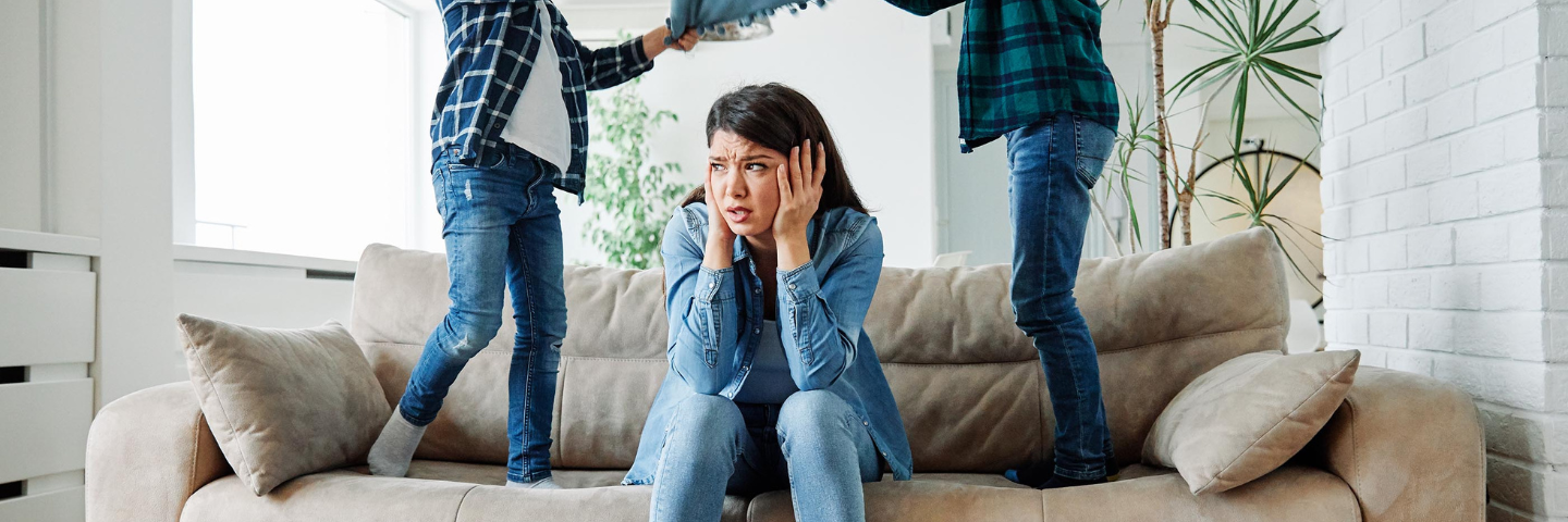 Une mère assise sur un sofa, visiblement dépassée, se tenant la tête entre les mains pendant que deux enfants sautent et crient autour d’elle dans le salon.