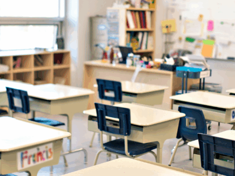 Salle de classe vide avec des bureaux et des chaises alignés, éclairée par de grandes fenêtres.