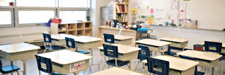 Salle de classe vide avec des bureaux et des chaises alignés, éclairée par de grandes fenêtres.