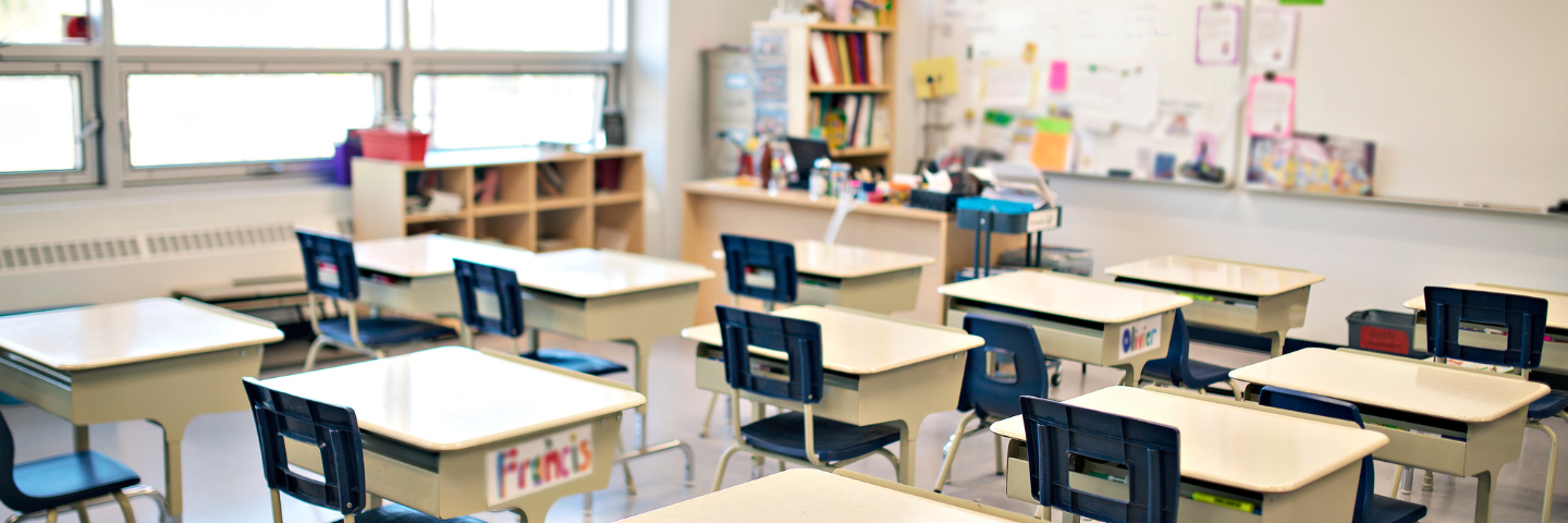 Salle de classe vide avec des bureaux et des chaises alignés, éclairée par de grandes fenêtres.