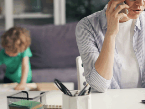 Parent en télétravail à la maison, en appel téléphonique devant un ordinateur portable, tandis qu’un jeune enfant joue ou fait une activité en arrière-plan.