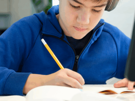 Jeune garçon concentré en train d’écrire dans un cahier à son bureau, avec un écran d’ordinateur à côté.