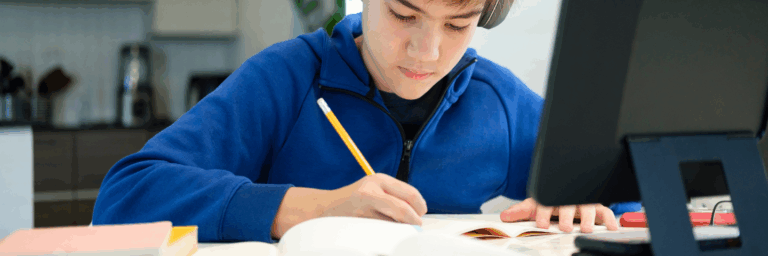 Jeune garçon concentré en train d’écrire dans un cahier à son bureau, avec un écran d’ordinateur à côté.